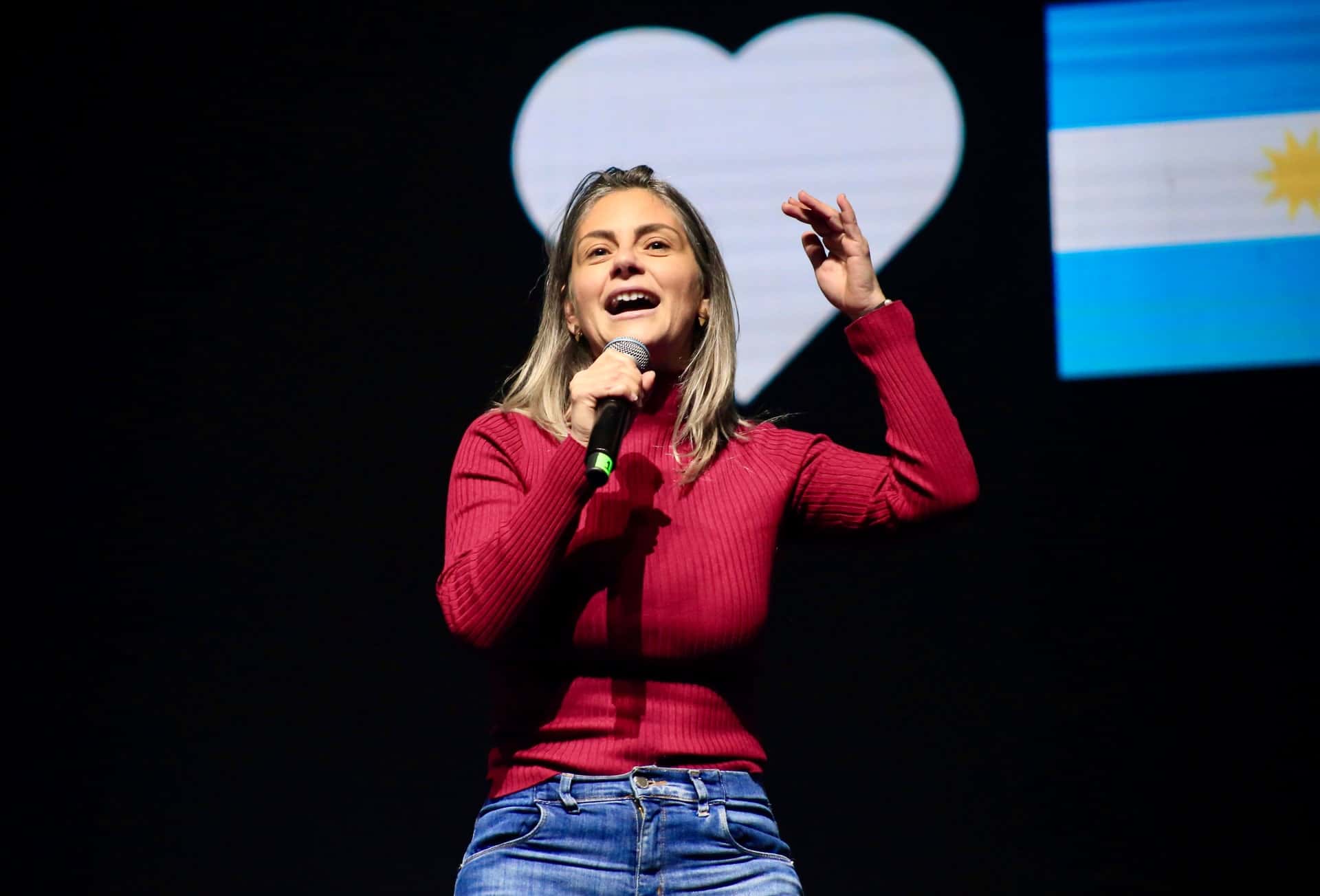 Micaela Ferraro habla en un escenario con una bandera argentina de fondo